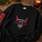 Folded black Permanently On The Naughty List Christmas jumper on a wooden surface with a tartan blanket and candle.