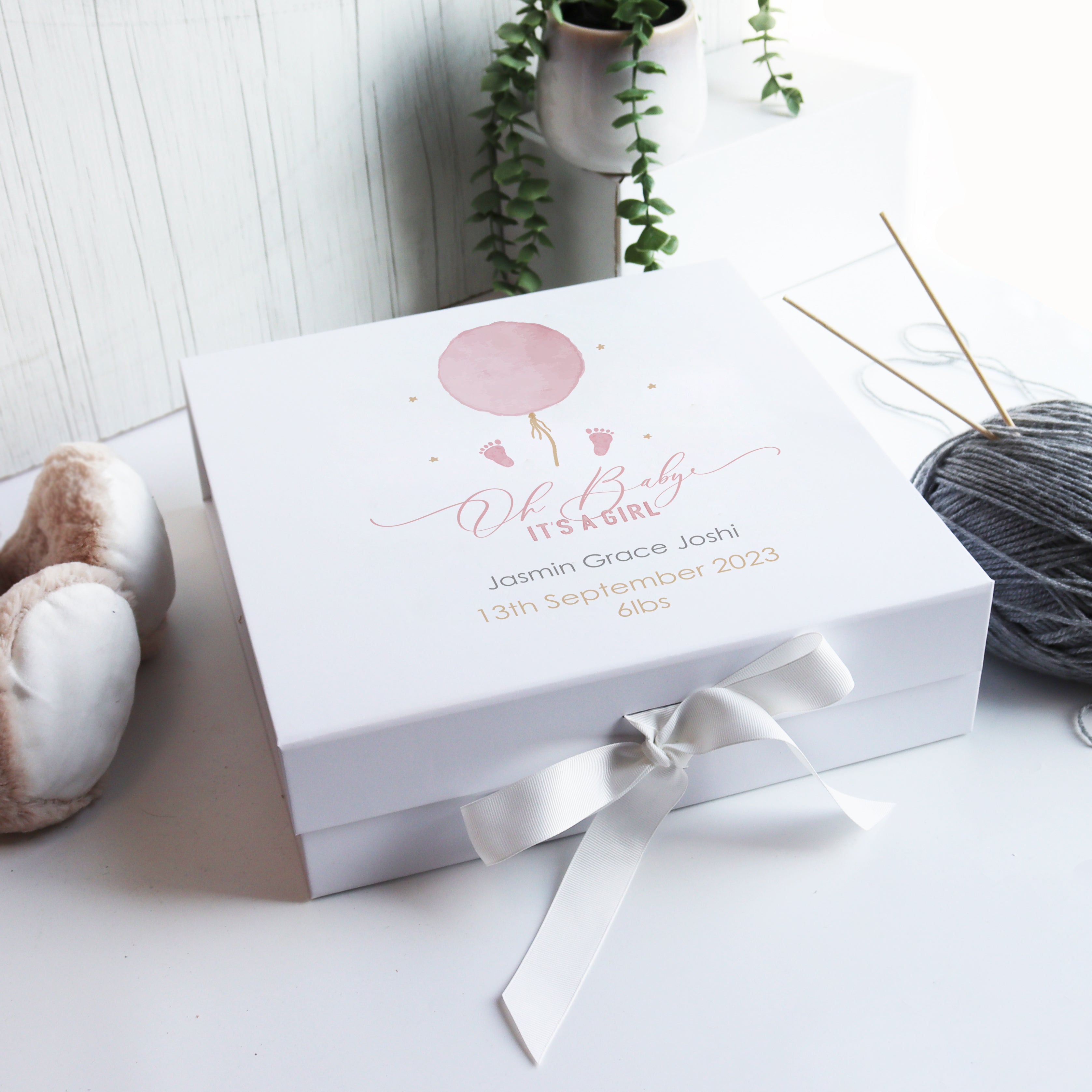 A white luxury baby gift box with a hand-drawn watercolour design of a single balloon, personalized with the baby's name and birth details, presented with a white ribbon and placed on a wooden surface with a plant and knitting items in the background.