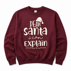A maroon crewneck sweatshirt featuring a white graphic print that reads 'DEAR Santa i can EXPLAIN' with a small Santa hat illustration above the text, displayed on a clean white background.
