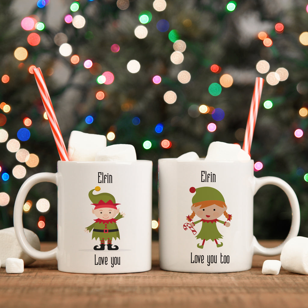 Two white ceramic mugs with 'Elfin Love You' and 'Elfin Love You Too' text, featuring illustrations of elves, placed on a table with Christmas decorations in the background.