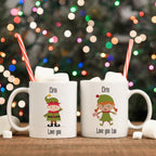 Two white ceramic mugs with 'Elfin Love You' and 'Elfin Love You Too' text, featuring illustrations of elves, placed on a table with Christmas decorations in the background.