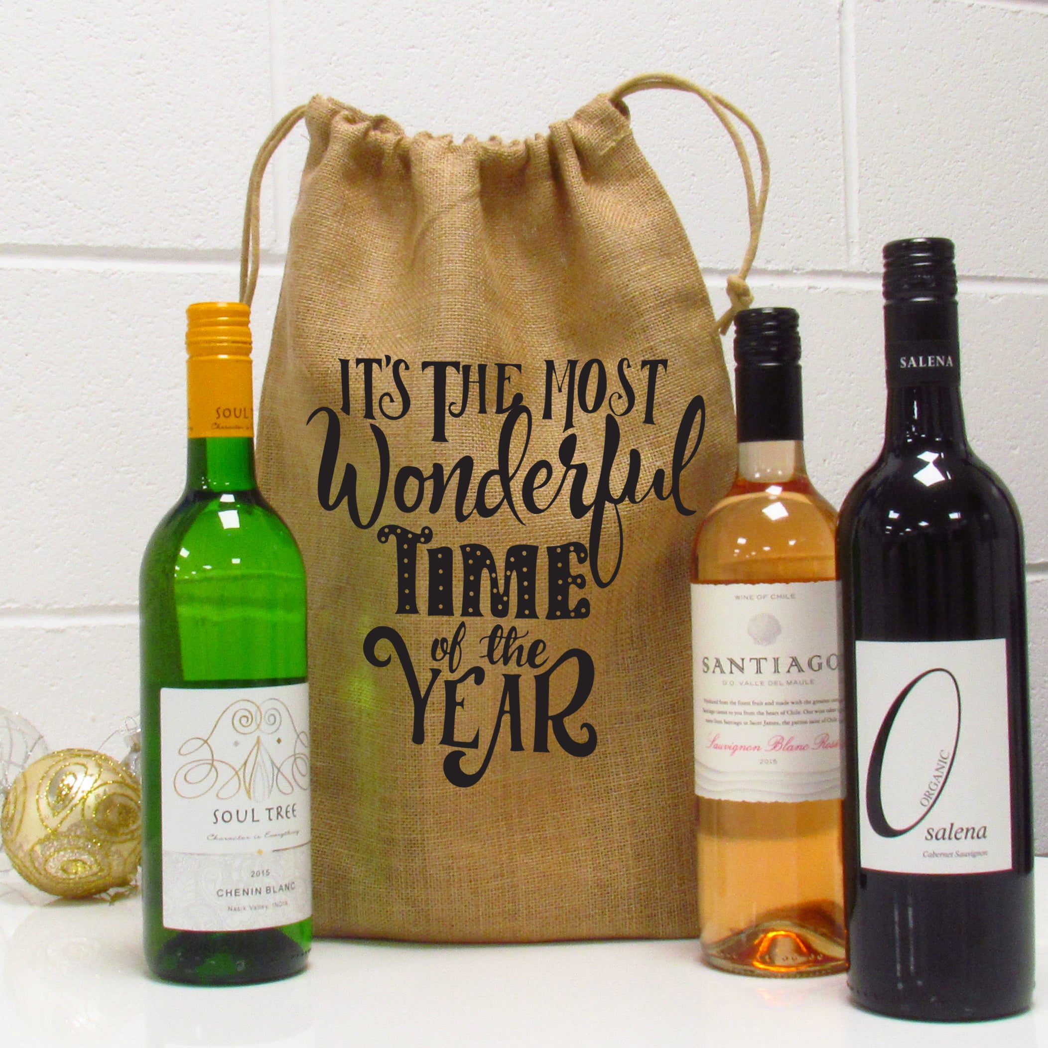 A burlap wine gift bag with 'It's The Most Wonderful Time Of The Year' text, displayed alongside three wine bottles.