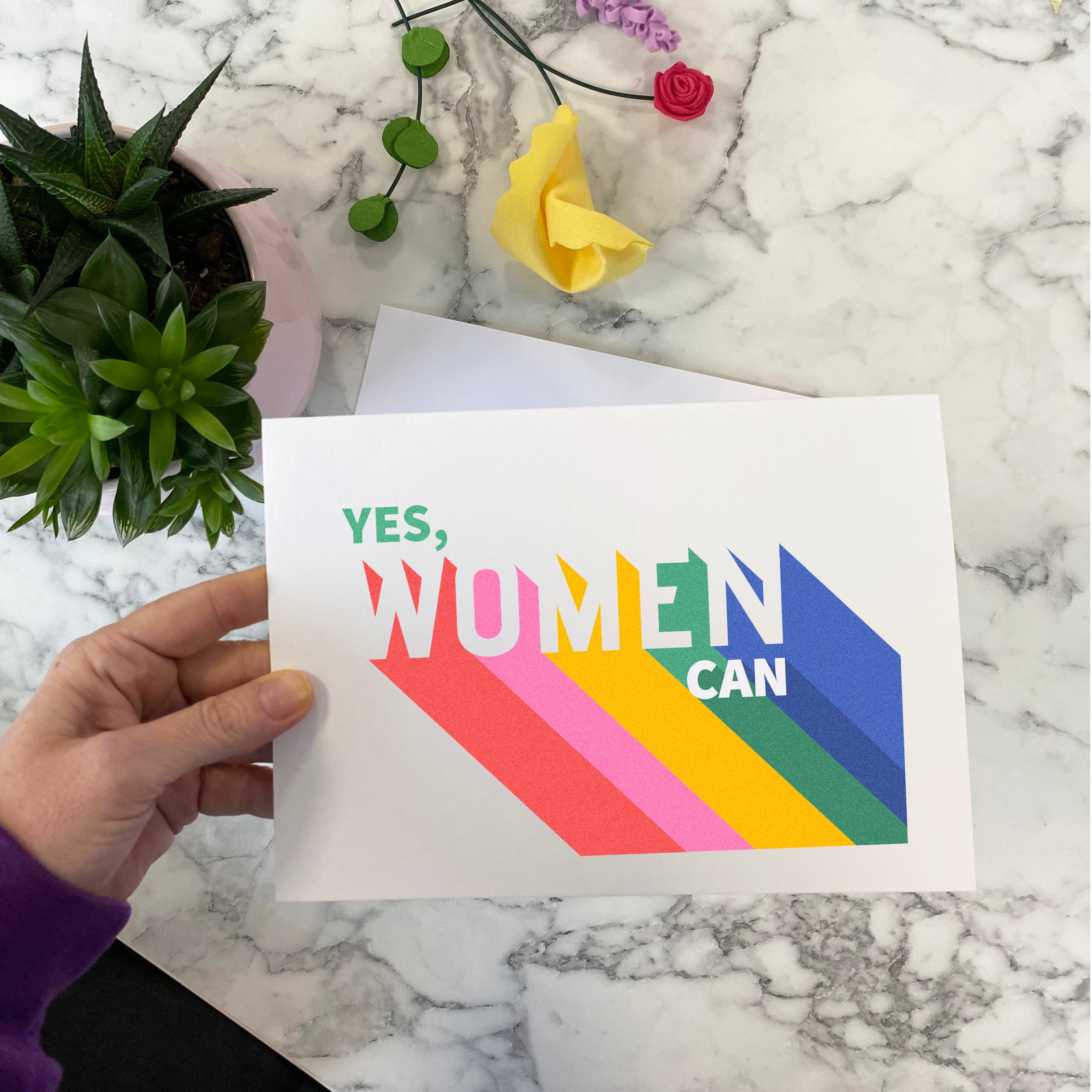 A colorful card with the text 'YES WOMEN CAN' in a geometric pattern, held by a hand against a backdrop of a potted plant and a customized greetings card.