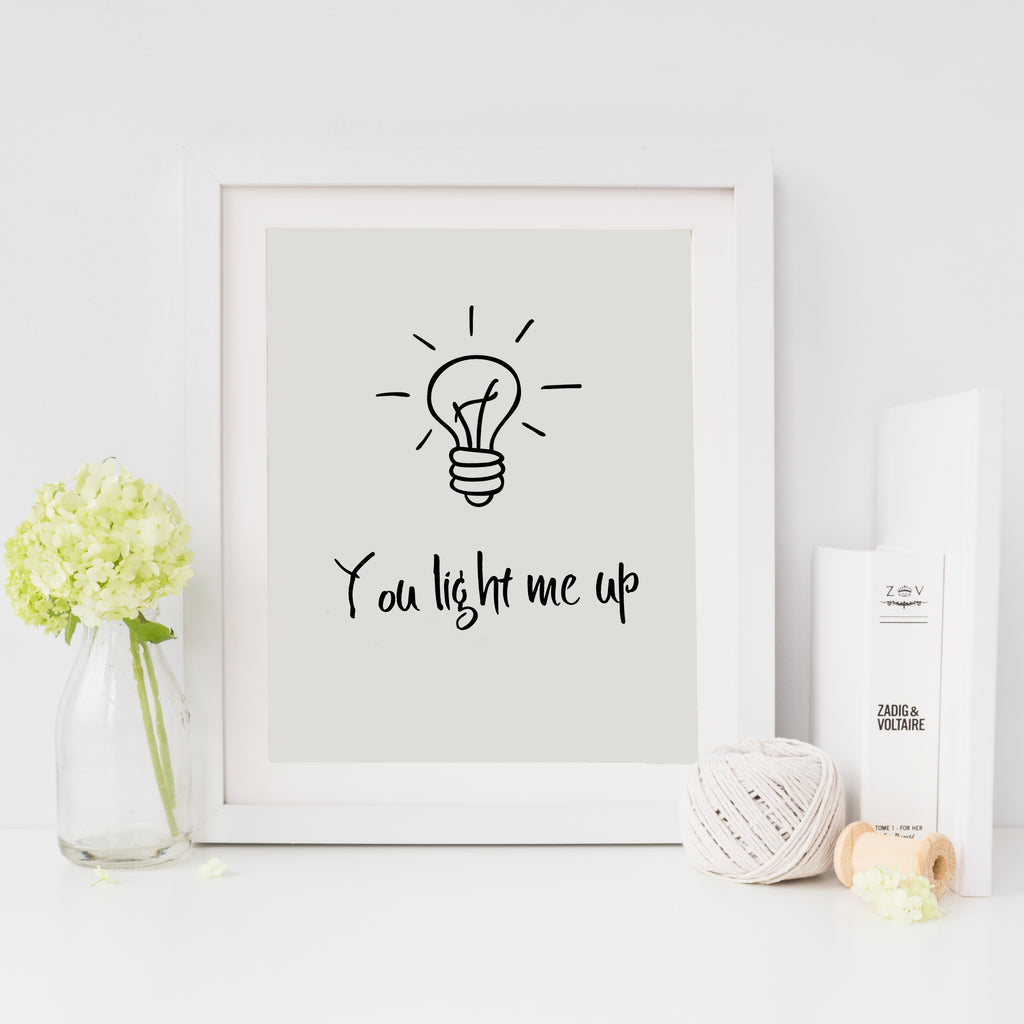 A contemporary print with a light bulb and the phrase 'you light me up' written on it, presented in a white or black frame, sitting on a table with decorative items around it.
