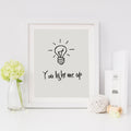 A contemporary print with a light bulb and the phrase 'you light me up' written on it, presented in a white or black frame, sitting on a table with decorative items around it.