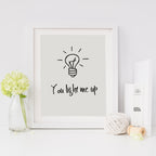A contemporary print with a light bulb and the phrase 'you light me up' written on it, presented in a white or black frame, sitting on a table with decorative items around it.