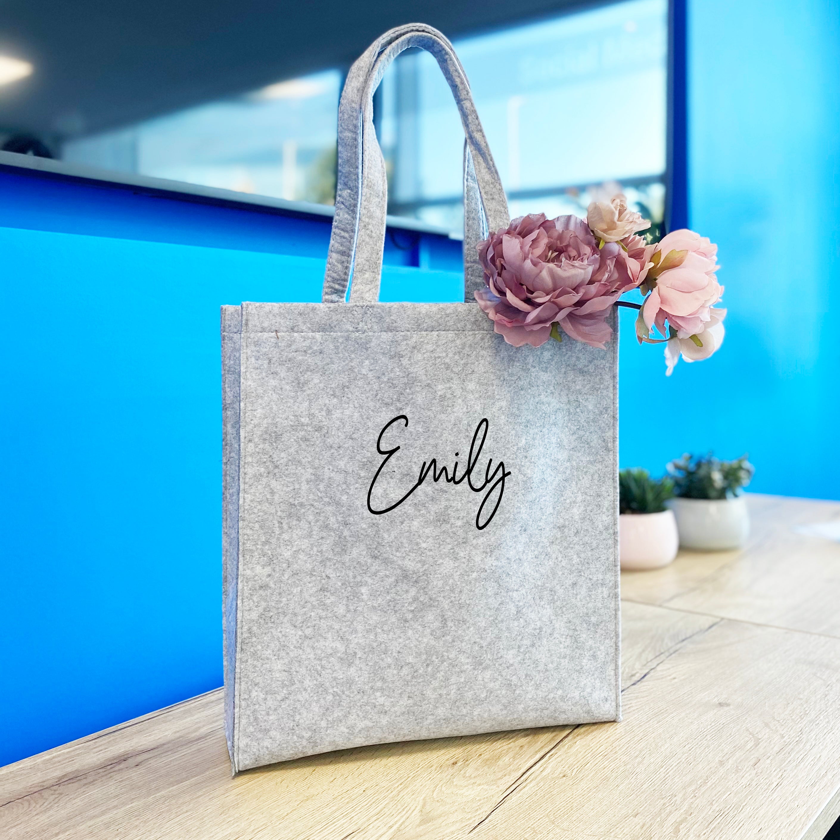 A personalised gray felt tote bag with a white print name 'Emily' on the front, placed on a table with a pink flower decoration and a blue background.