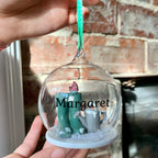 A personalised glass bauble with a green and white color scheme, containing a figurine of a pair of gardening gloves, and the name 'Margaret' written on it.