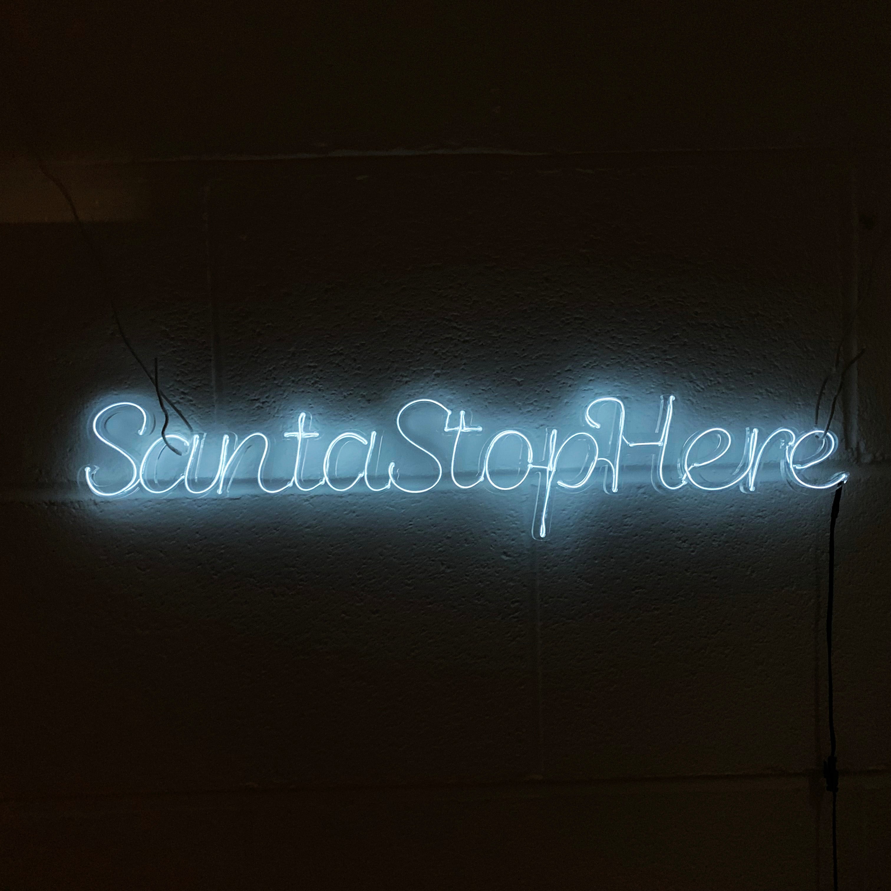 Neon sign with the text 'Santa Stop Here' written in blue, mounted on a wall.