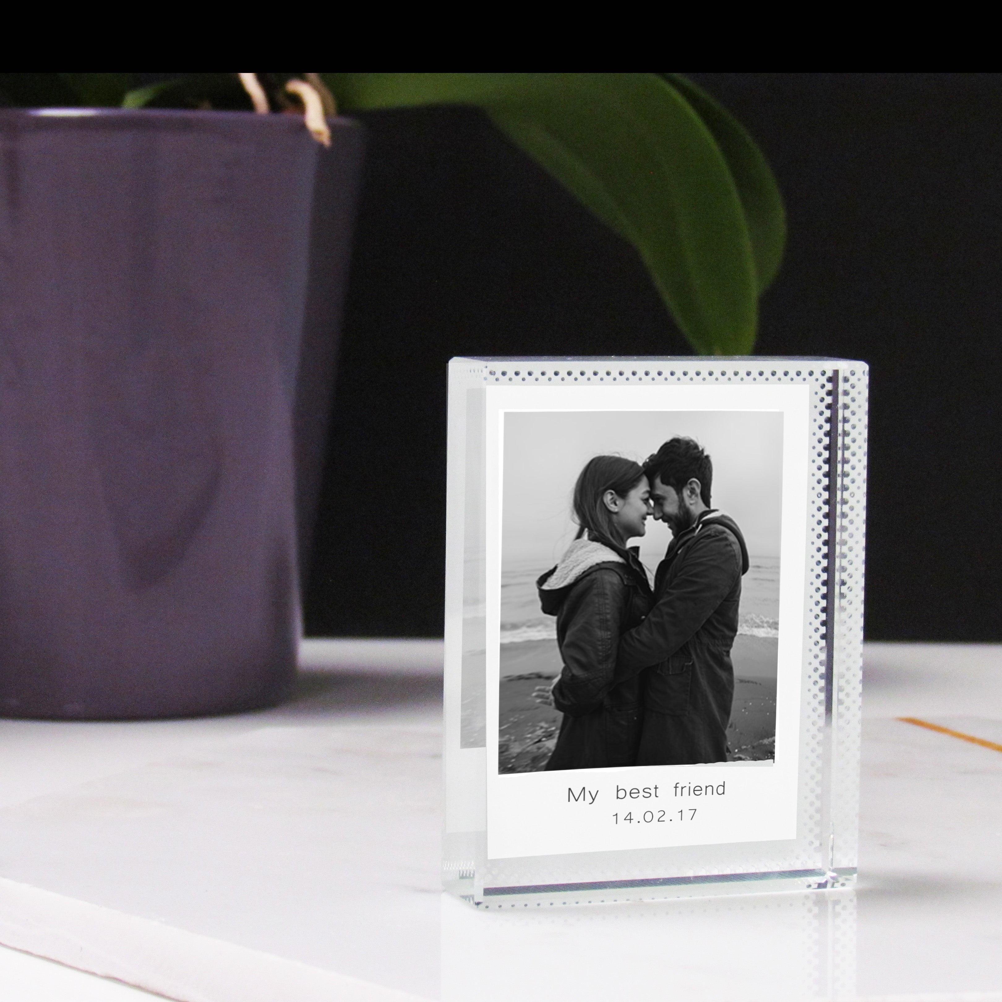 A clear acrylic photo block with a couple embracing printed on it, with a white background, placed on a marble surface next to a potted plant.