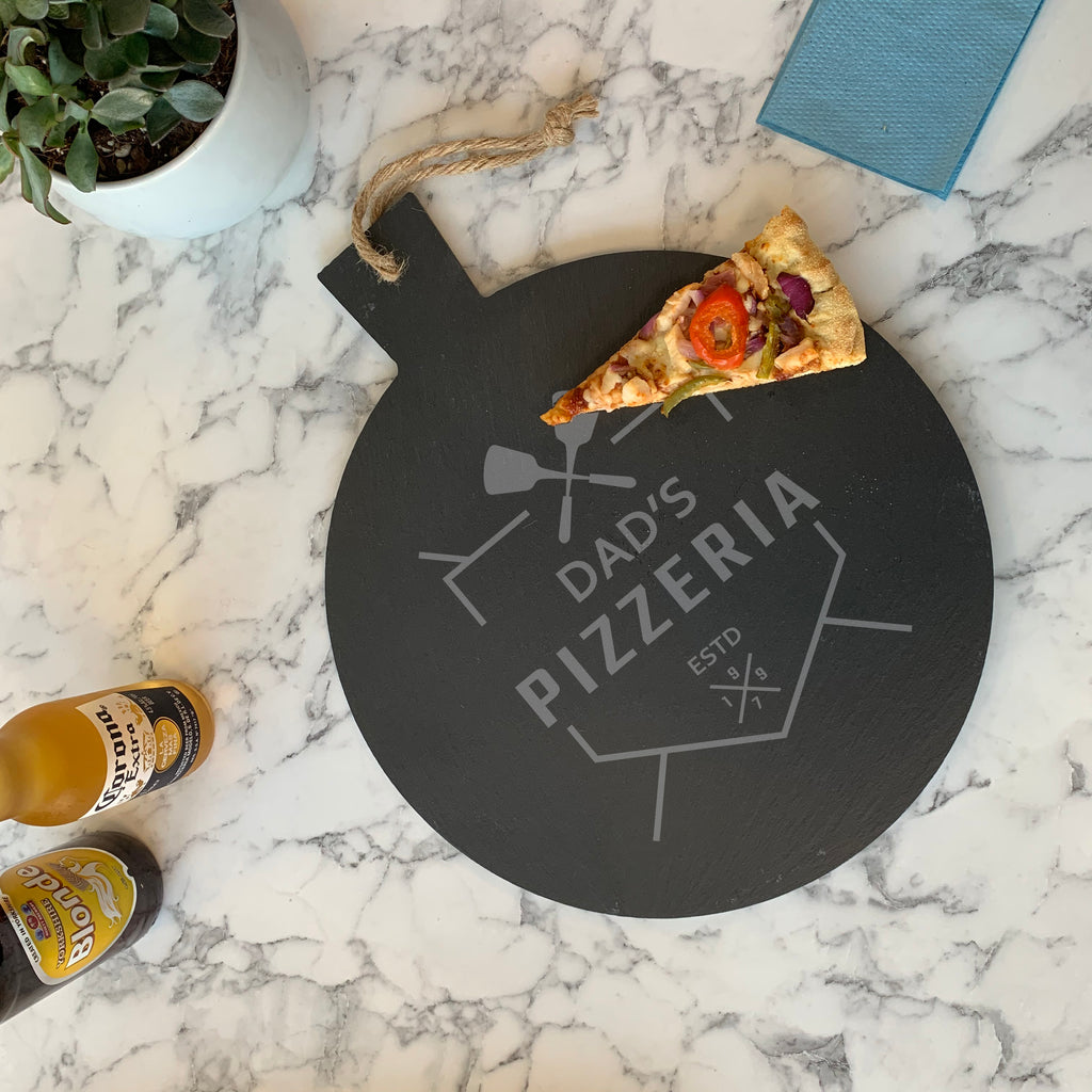 A round slate pizza board with 'Dad's Pizza' text and a pizza slice cut out for display. The board is black with a grayish appearance, and there are some beverage bottles and a plant in the background.