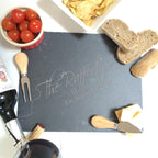 A personalized engraved slate cheeseboard with a knife, bread, tomatoes, and a bottle of wine in the background, on a white surface.