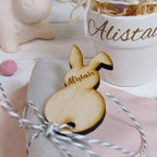 A wooden Easter bunny-shaped napkin holder with a name 'Alastair' engraved on it, placed on a pink cloth, next to a white cup and a decorative branch.