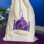 A white gift sack with a purple and gold glitter design, featuring a graphic of a mosque and minaret, and the word 'Eid' with a personalized name below.