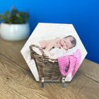 A hexagonal photo gift tile with an image of a baby in a basket, placed on a wooden surface with a blue background.