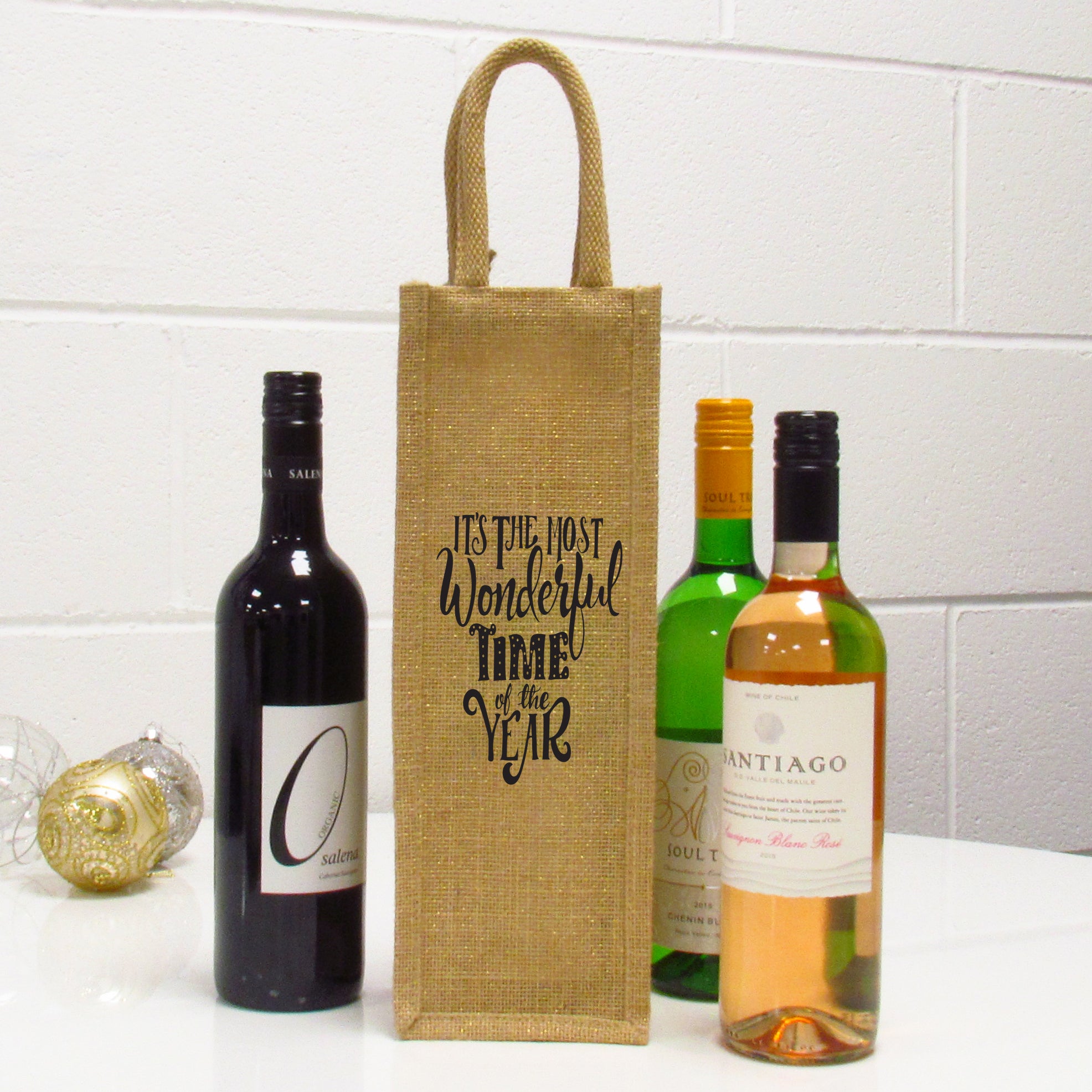 A beige jute wine bag with a Christmas-themed message 'It's The Most Wonderful Time Of The Year' printed in black. The bag is fitted with a bottle, and stands upright next to two wine bottles.