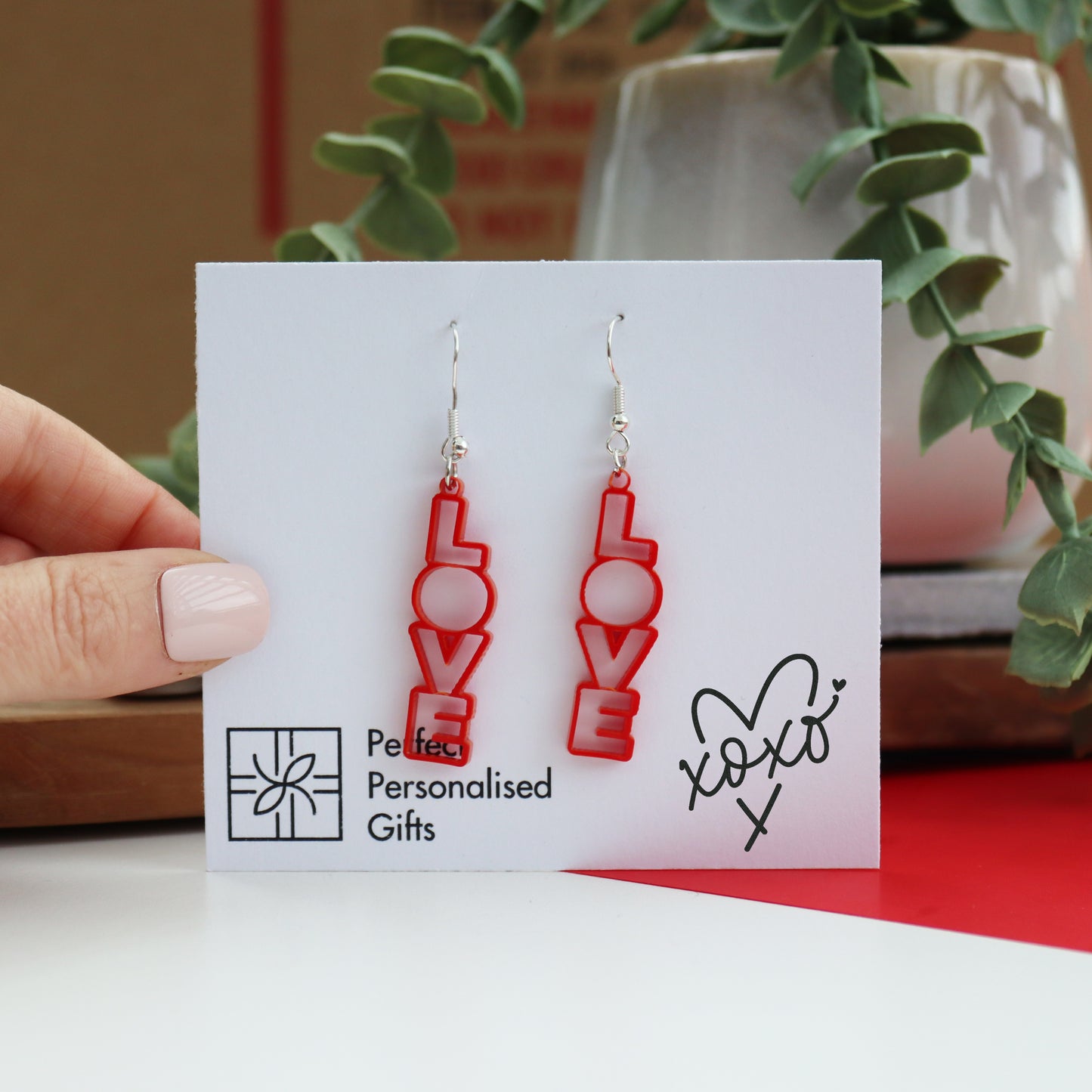 Love drop earrings cut from red acrylic on a white backing card with a xoxo design printed on the left bottom corner and the brand printed on the right bottom corner