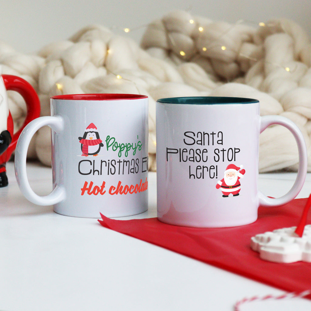 Two ceramic mugs, one with a red and white design reading 'Santa's Please Stop Here!' and the other with a green and white design reading 'Emily's Christmas Eve Hot Chocolate'. Both mugs have Christmas-themed decorations and are placed on a white surface with a red blanket and Christmas lights in the background.