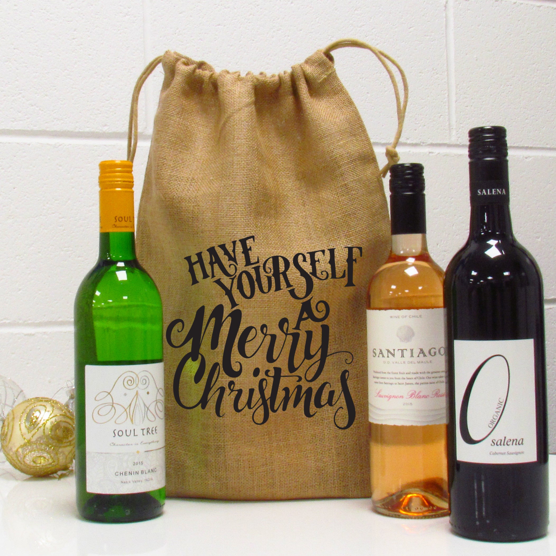 A burlap wine gift bag with 'Have Yourself A Merry Christmas' text printed on it, alongside three wine bottles.