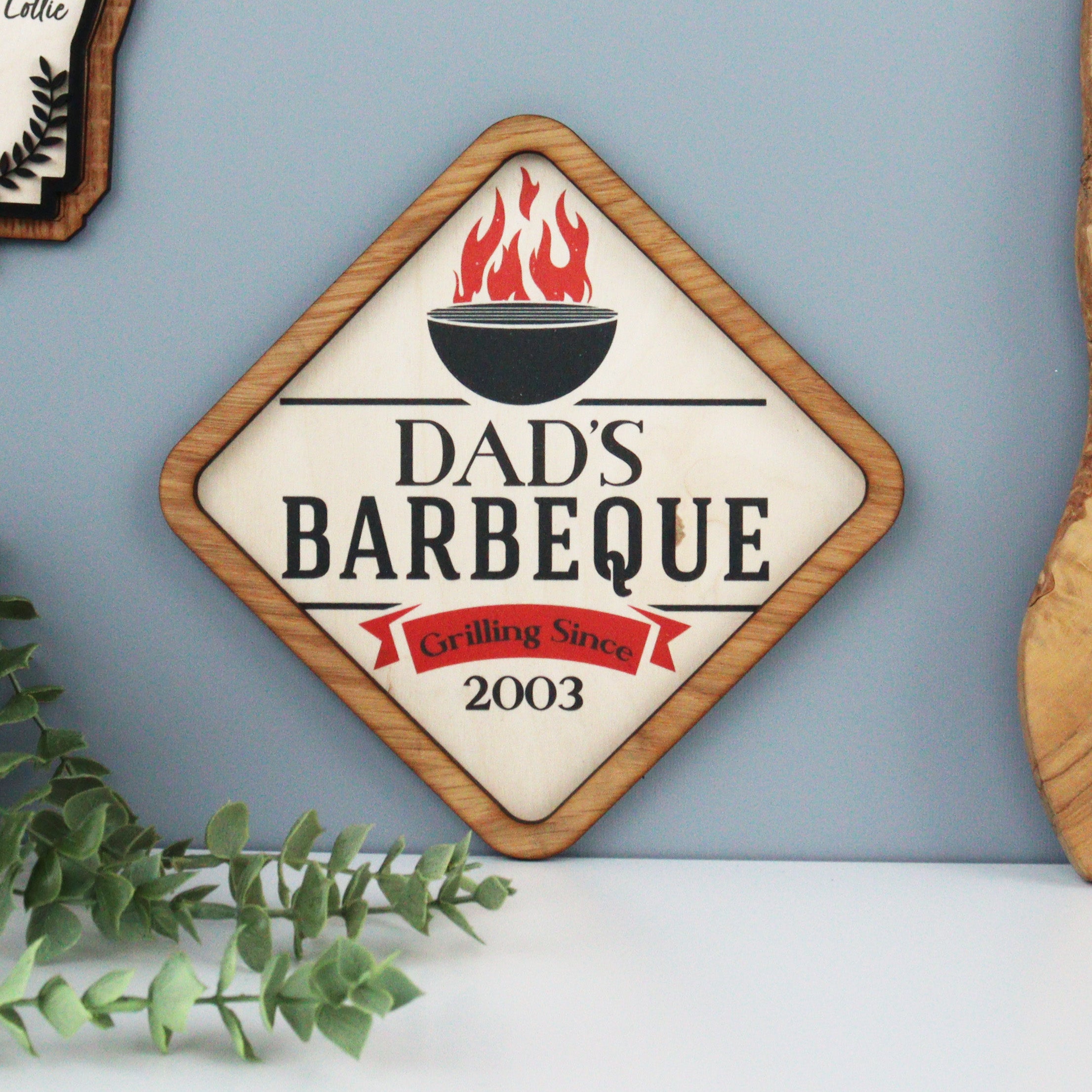 A personalised barbecue sign plaque with 'Dad's Barbecue' text, a bowl with a flame on top, and a custom name and date below the text.