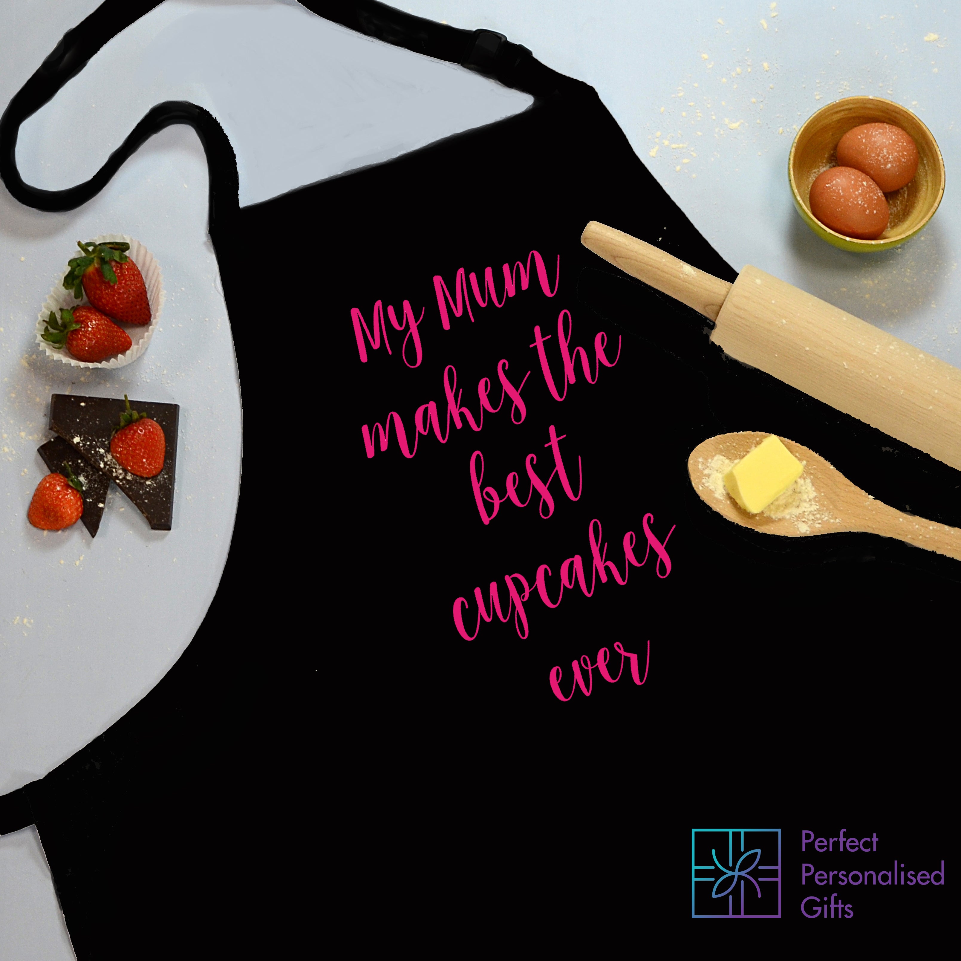 A black apron with a personalized message that reads 'MY MUM THE BEST CUPCAKES EVER' along with an image of a wooden spoon and some cupcakes. The apron is hanging on a hook with a blue and white brand tag 'PERFECT PERSONALIZED GIFTS'.