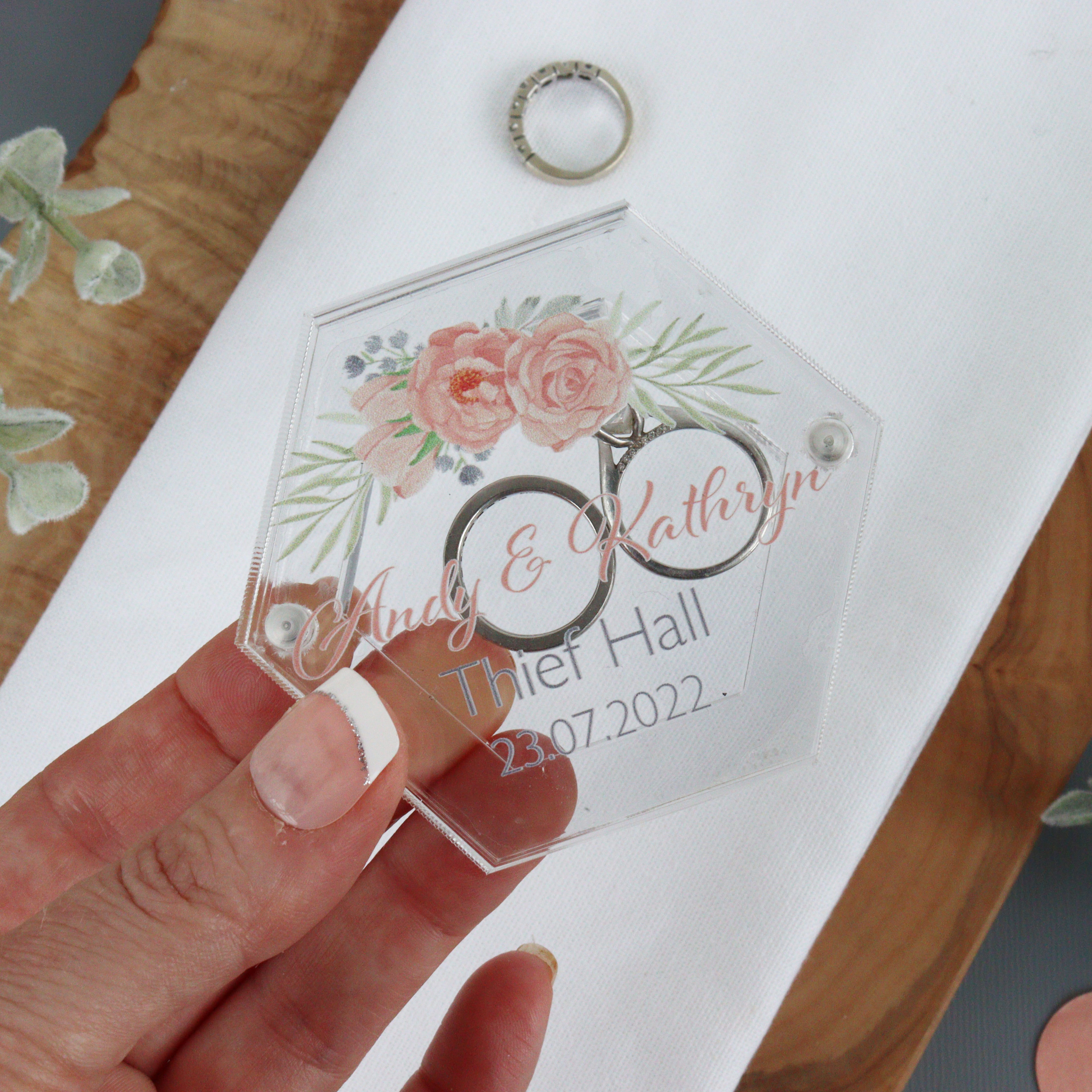A clear acrylic personalised wedding ring box being held by a hand, with a ring placed inside it. The box is hexagonal in shape and has printed text including names and a date.