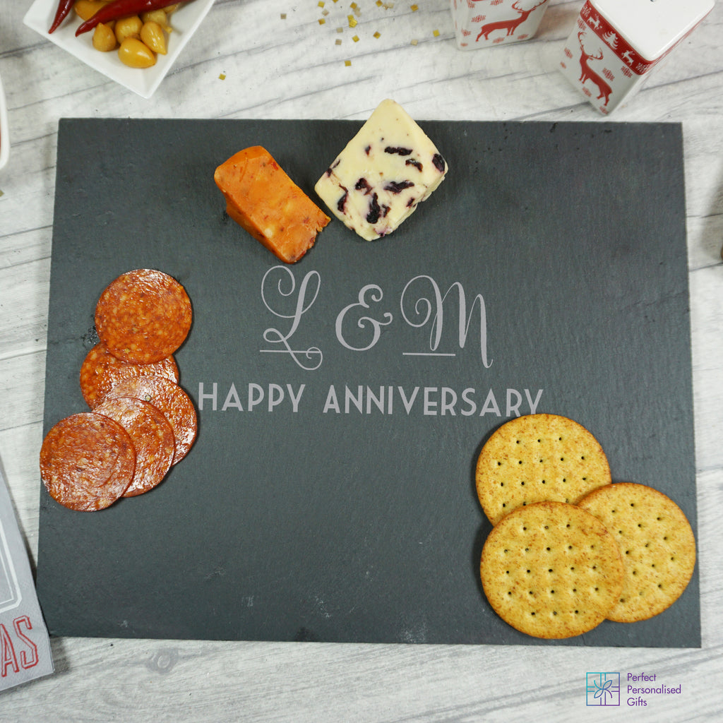 A personalized slate cheeseboard with the text 'L&M' and 'Happy Anniversary' on it, accompanied by sliced cheese and crackers.