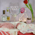 An engraved clear glass gin goblet with a text 'MUM'S G&T' on a table, with decorative items and a pink cloth in the background.