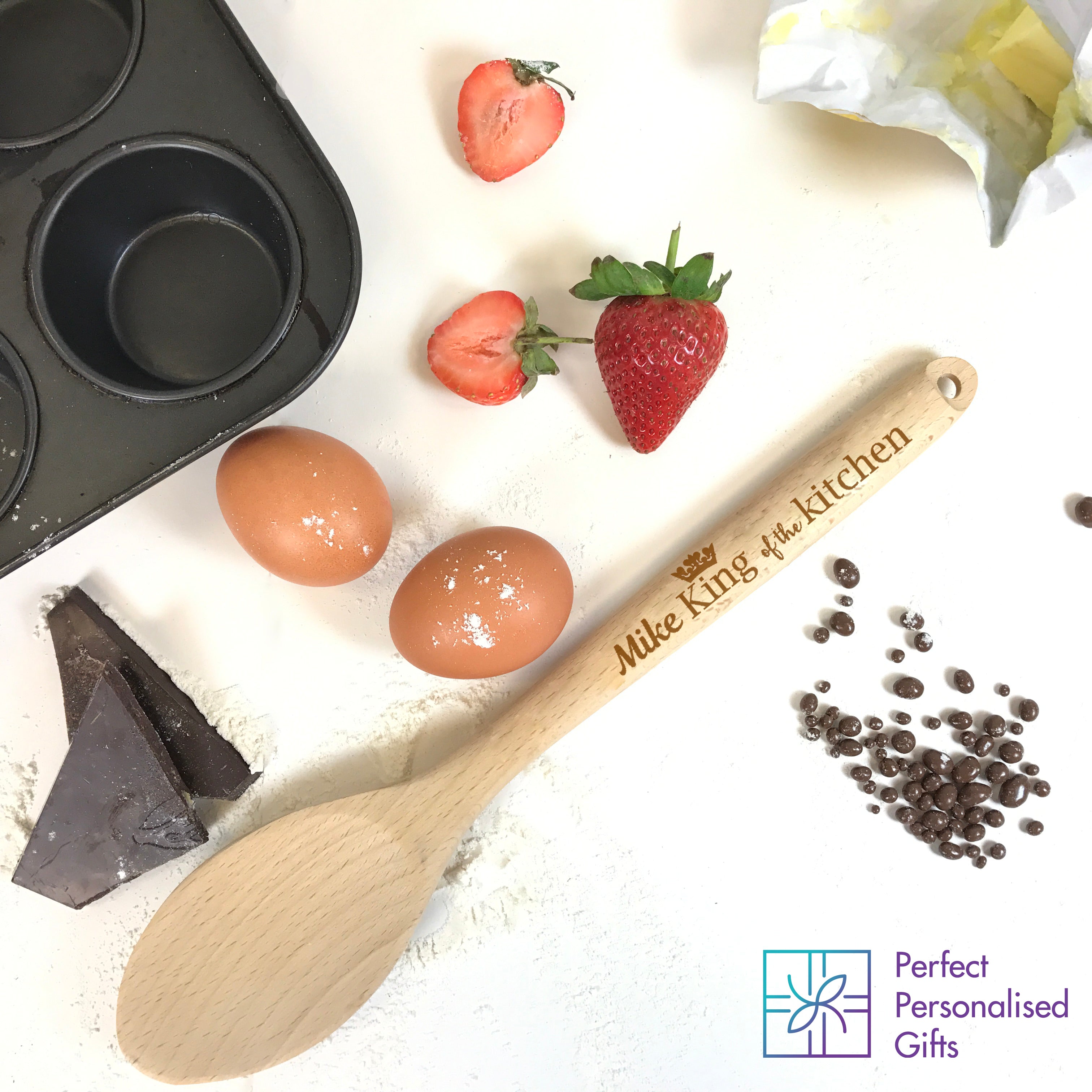 A wooden spoon with the engraved text 'King of the Kitchen' along with a name, in this case 'Mike', on a wooden surface with ingredients like eggs, strawberries, and a bar of chocolate in the background, suggesting a kitchen setting.