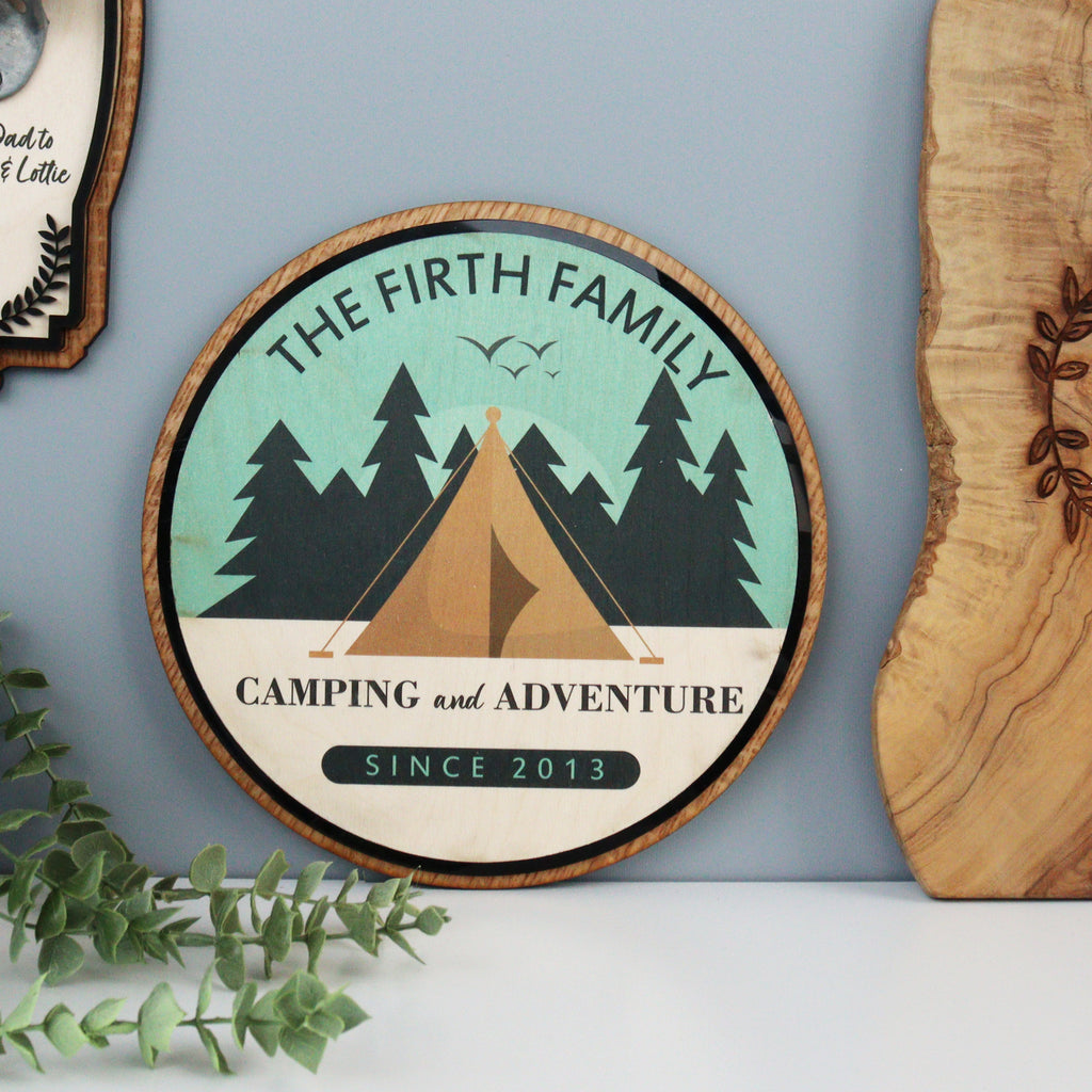 A personalised wooden sign with a green and brown color scheme, featuring text and graphics related to camping, placed on a wooden surface.