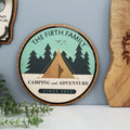 A personalised wooden sign with a green and brown color scheme, featuring text and graphics related to camping, placed on a wooden surface.