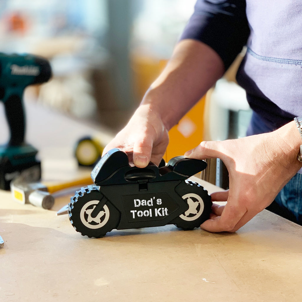 A person is holding a black motorcycle-shaped tool kit with the text 'Dad's Tool Kit' on it. The kit is presented with various tools such as wrenches and pliers, arranged in a workshop setting.