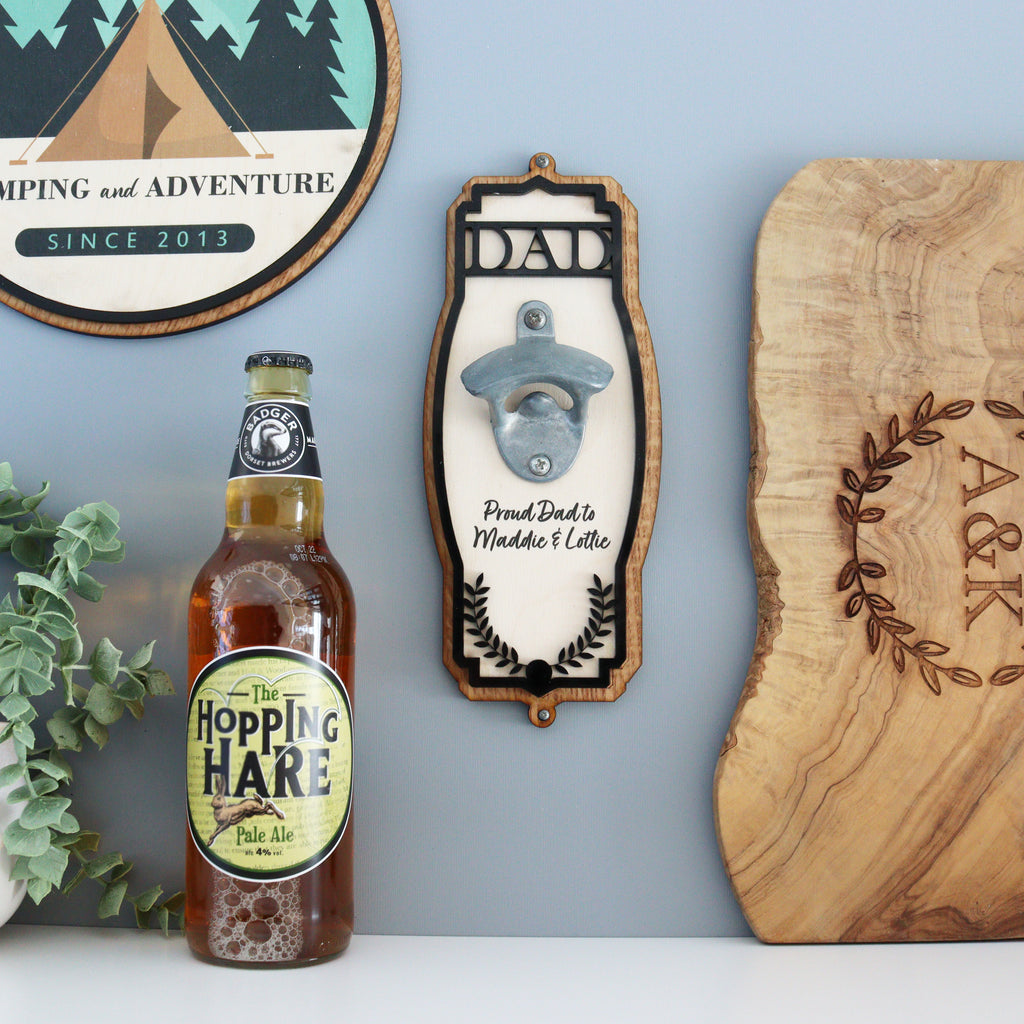 A wall-mounted personalised bottle opener with a wooden appearance and black ornate acrylic trim, featuring a stainless steel vintage-style bottle opener in the center. The opener has 'DAD' engraved on the front, with a round plaque above it that reads 'PROUD DAD CUTS DEEP', and a round ornate acrylic piece above that with a tree and leaves motif.