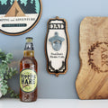 A wall-mounted personalised bottle opener with a wooden appearance and black ornate acrylic trim, featuring a stainless steel vintage-style bottle opener in the center. The opener has 'DAD' engraved on the front, with a round plaque above it that reads 'PROUD DAD CUTS DEEP', and a round ornate acrylic piece above that with a tree and leaves motif.