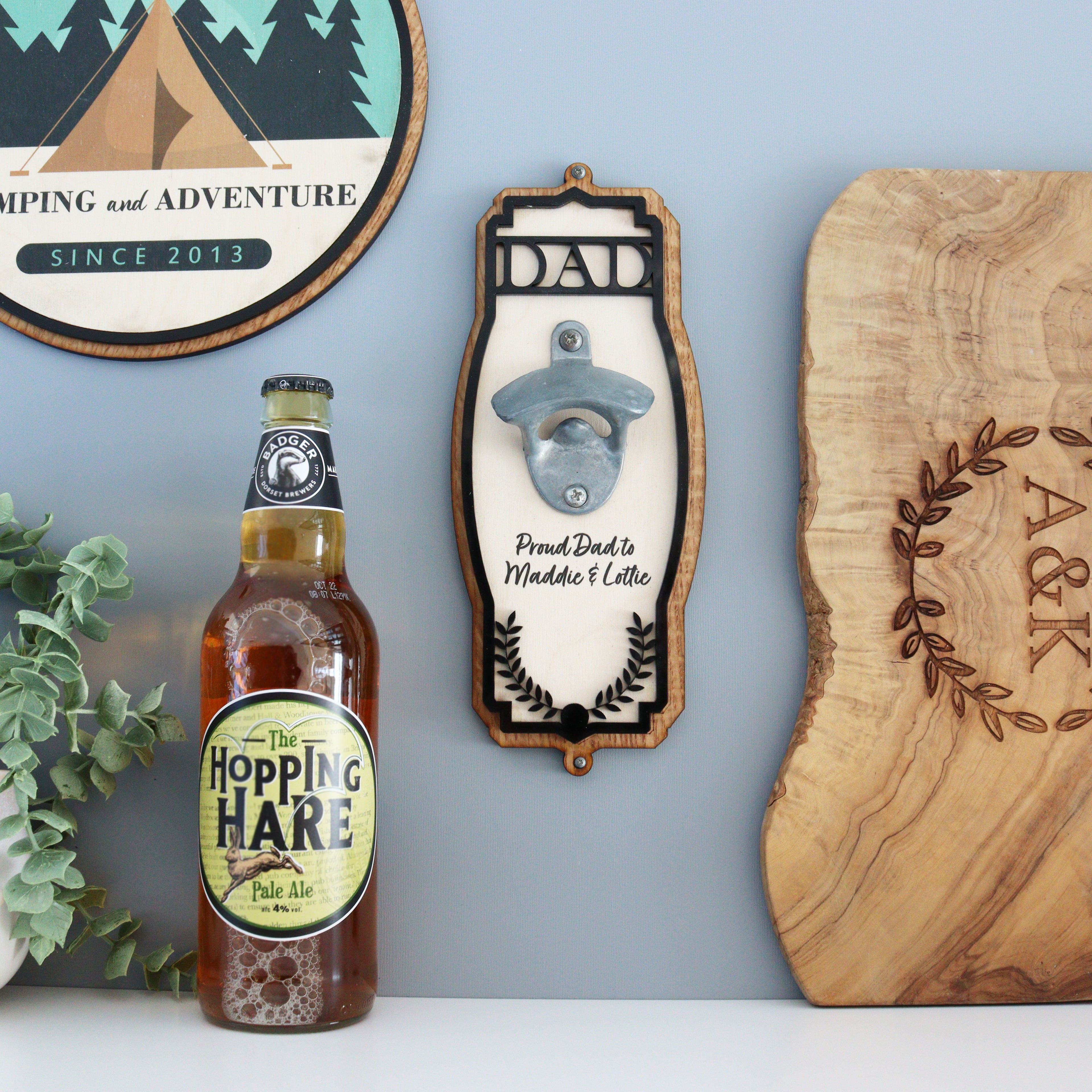 A wall-mounted personalised bottle opener with a wooden appearance and black ornate acrylic trim, featuring a stainless steel vintage-style bottle opener in the center. The opener has 'DAD' engraved on the front, with a round plaque above it that reads 'PROUD DAD CUTS DEEP', and a round ornate acrylic piece above that with a tree and leaves motif.