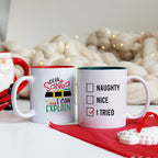 Two ceramic mugs, one with a red interior and the other with a green interior, featuring text and a tick box design on the front, placed on a surface with a Christmas-themed decoration.