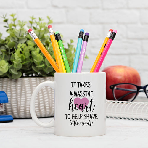 A white ceramic mug with a printed message that reads 'It takes a massive heart to help shape little minds!' alongside a colorful collection of pencils. The mug is presented on a table with a plant and glasses, indicating a personal touch and a humorous tone.