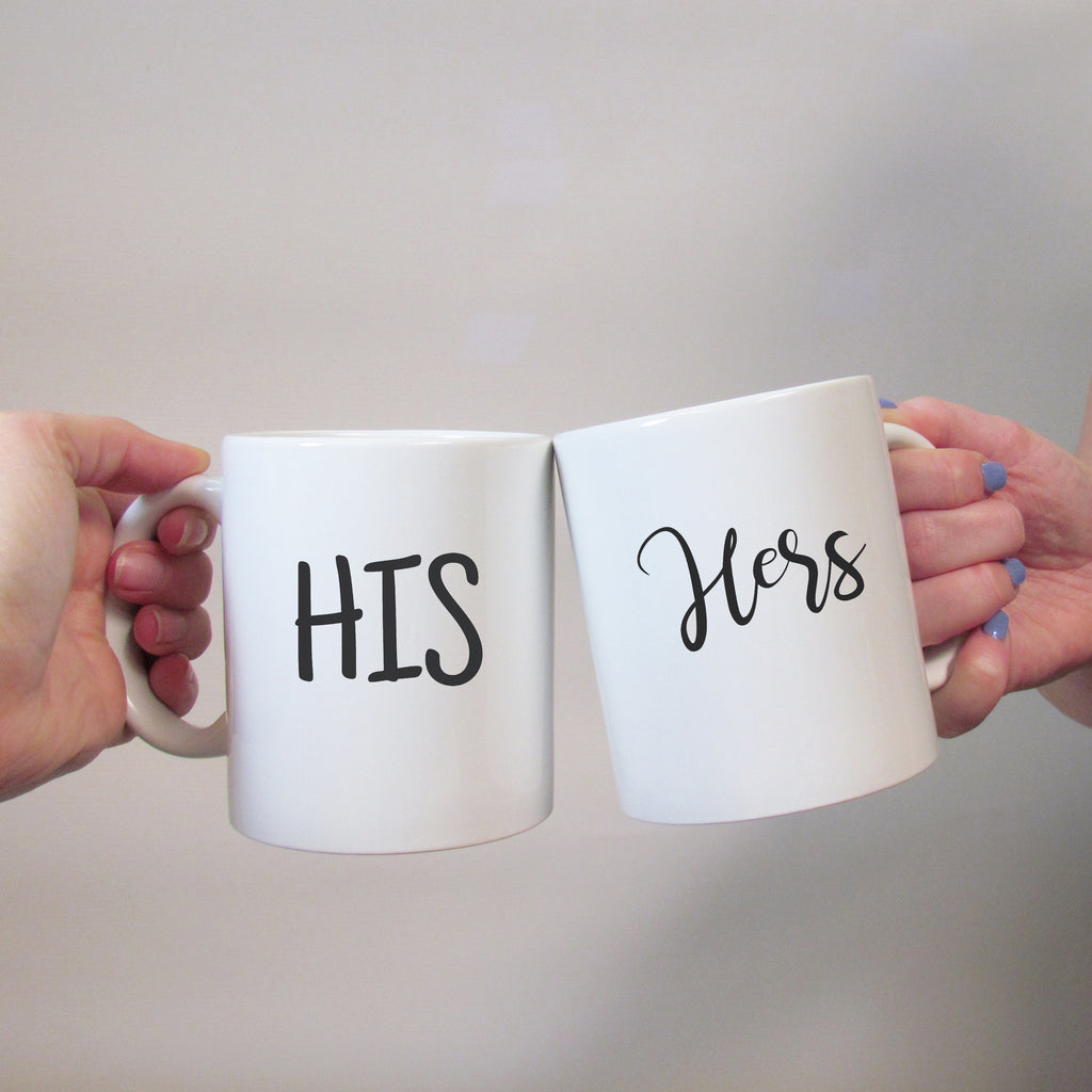 Two white ceramic mugs with the words 'HIS' and 'HERS' written in black script, held in hands against a neutral background.