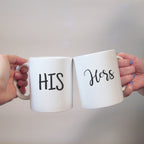 Two white ceramic mugs with the words 'HIS' and 'HERS' written in black script, held in hands against a neutral background.