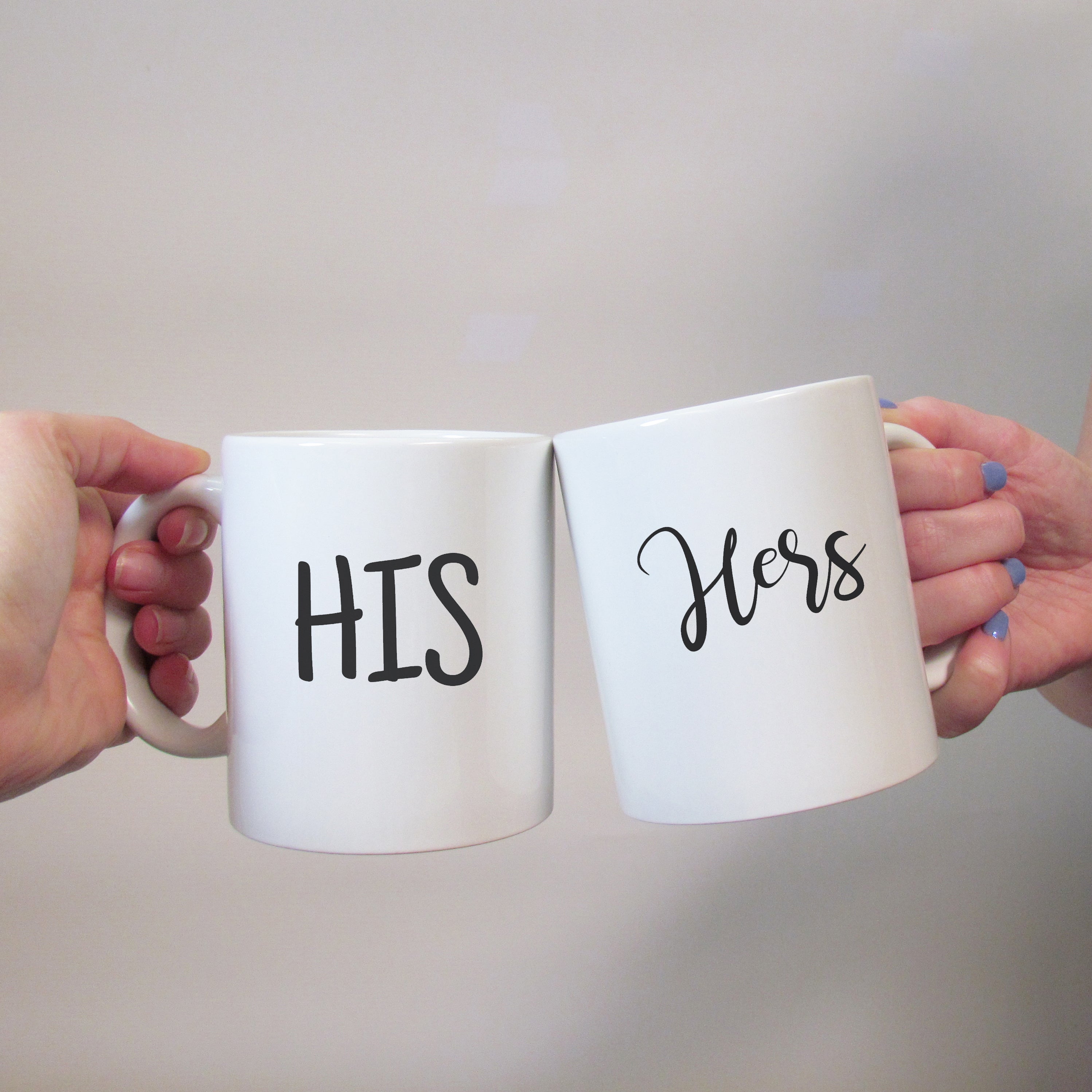 Two white ceramic mugs with the words 'HIS' and 'HERS' written in black script, held in hands against a neutral background.