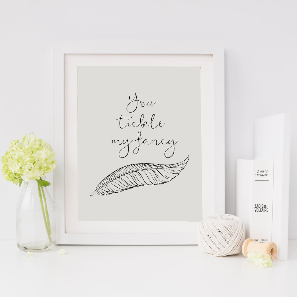 A minimalist art print with the phrase 'you tickle my fancy' and a feather illustration, in a grayscale color palette, framed and presented with decorative items around it.