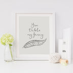 A minimalist art print with the phrase 'you tickle my fancy' and a feather illustration, in a grayscale color palette, framed and presented with decorative items around it.