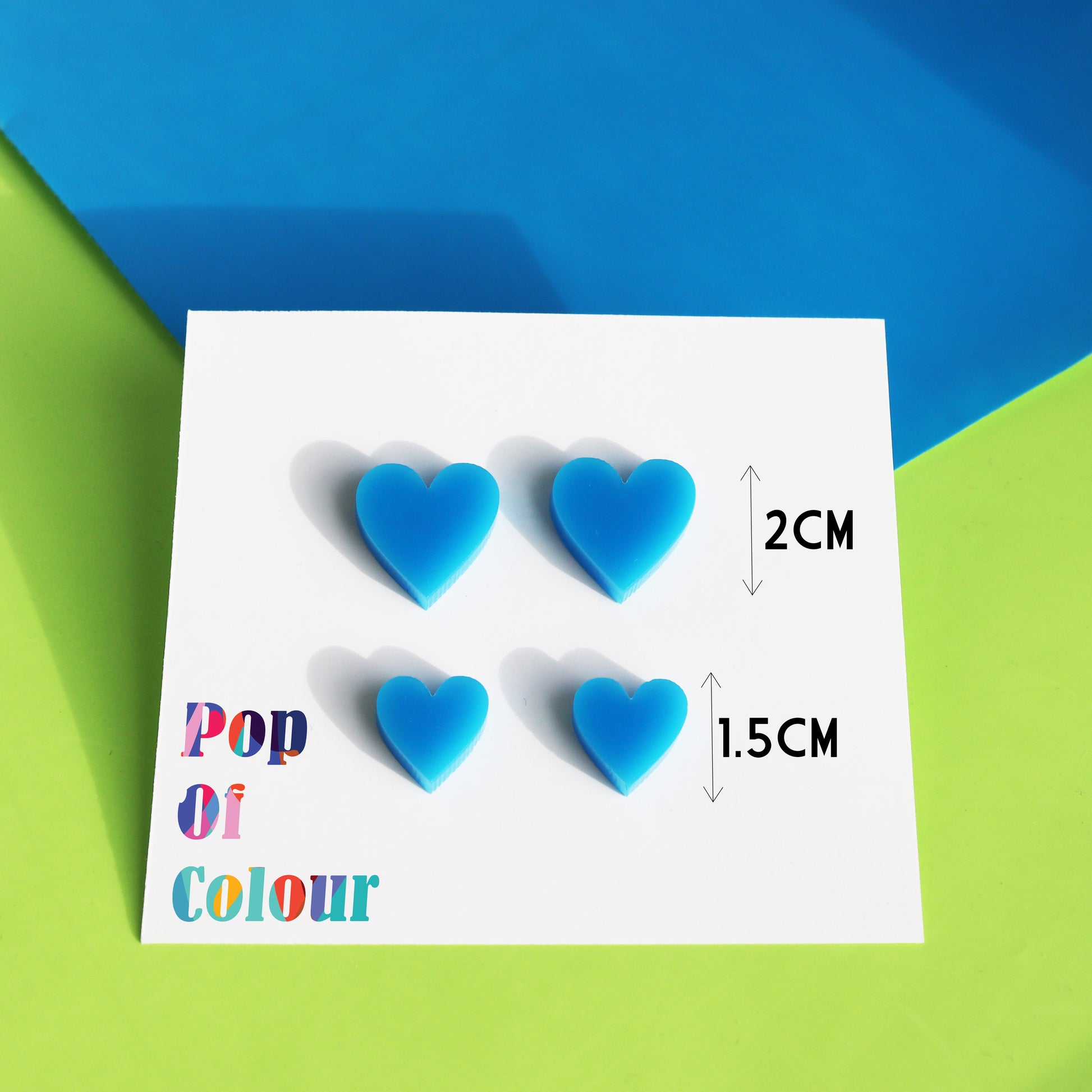 acrylic blue stud heart earrings available in two sizes shown on white earring backing card with Pop of Colour Logo in sunlight with sizes