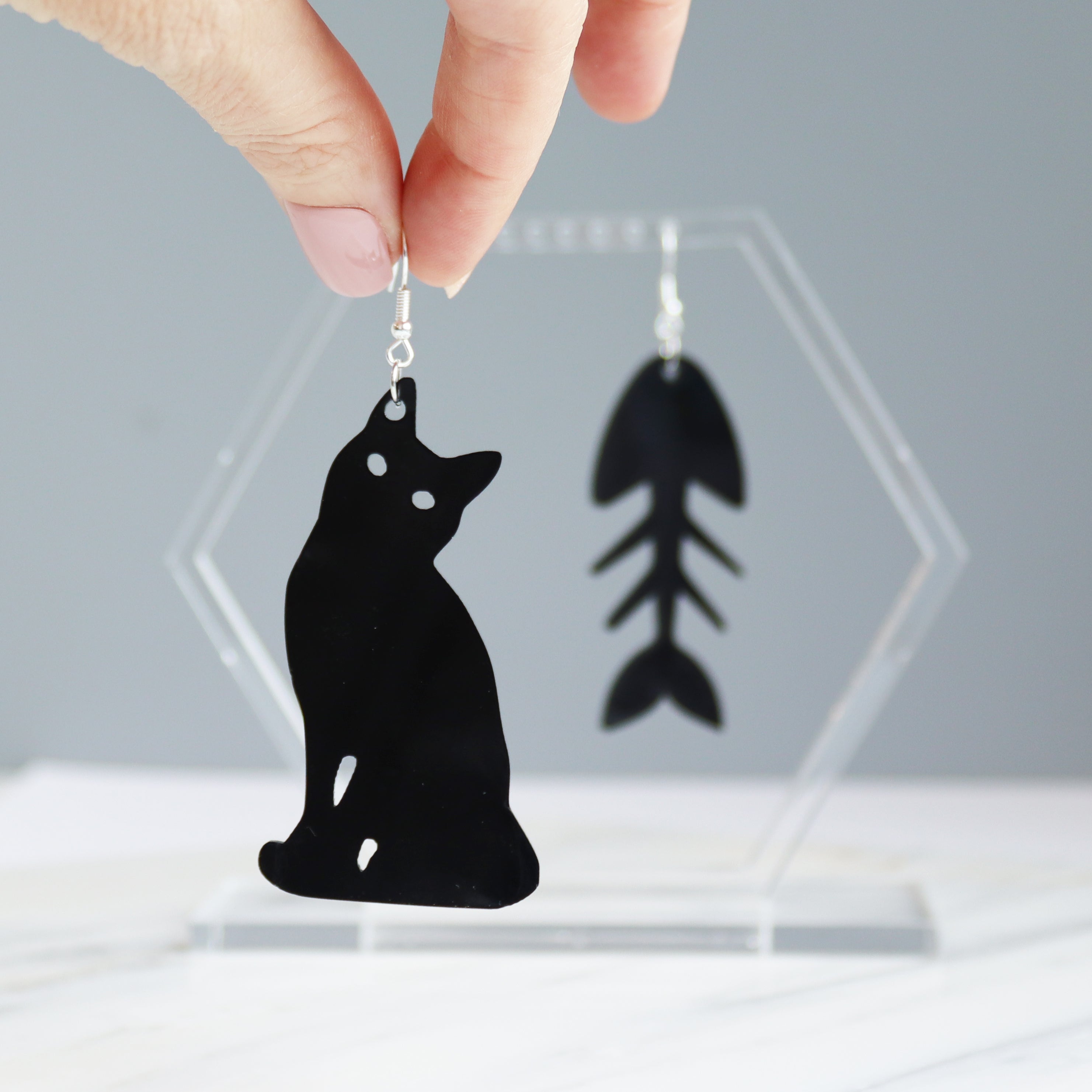 black cat and fish bone mismatches earrings cut from black acrylic with the bone earring on display stand on black cat shown being held by womans fingers