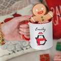 A ceramic mug with a Christmas character design, personalized with the name 'Emily'. The mug is held by a hand, and there are gifts and a red and white checkered blanket in the background.