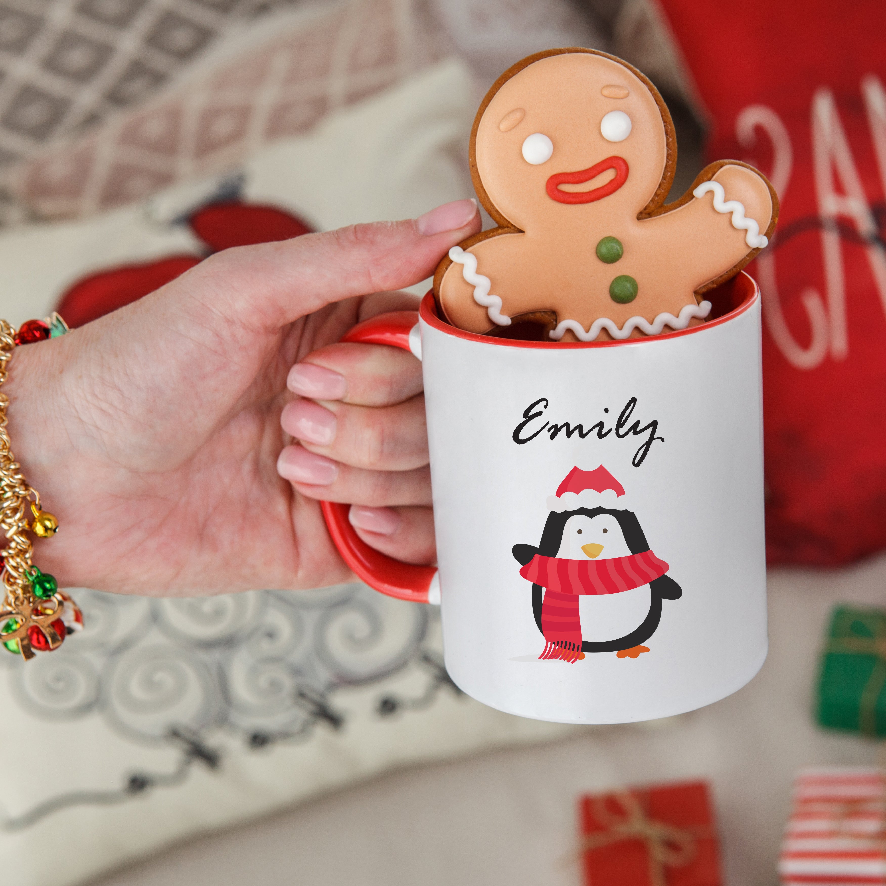A ceramic mug with a Christmas character design, personalized with the name 'Emily'. The mug is held by a hand, and there are gifts and a red and white checkered blanket in the background.