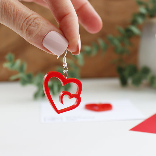 close up of bold red acrylic swirl heart earrings which are shown hanging from a persons hand