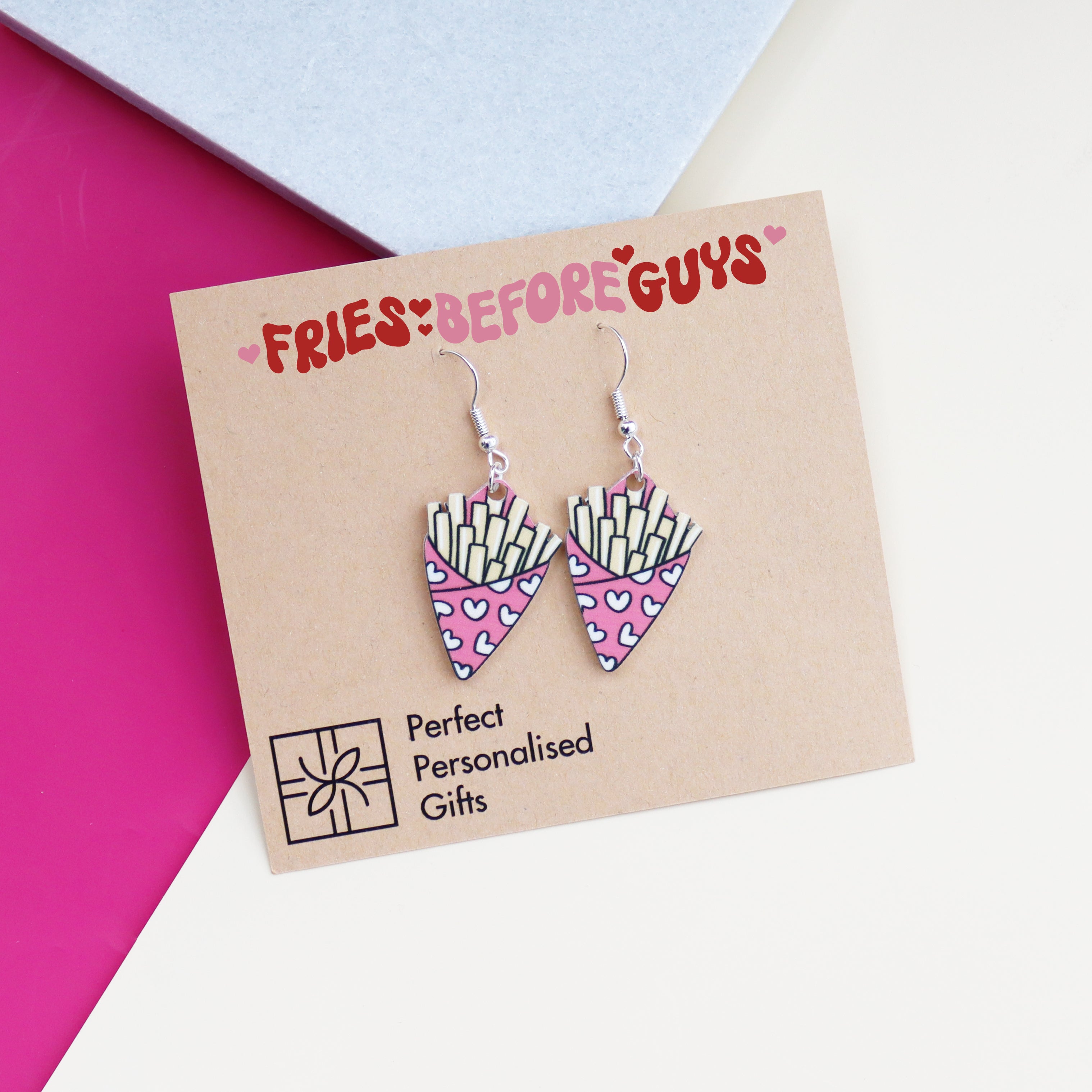 fries before guys earrings cut from acrylic and printed with a bag of chips design the bag of chips is pink and has hearts printed on it shown with an arty background