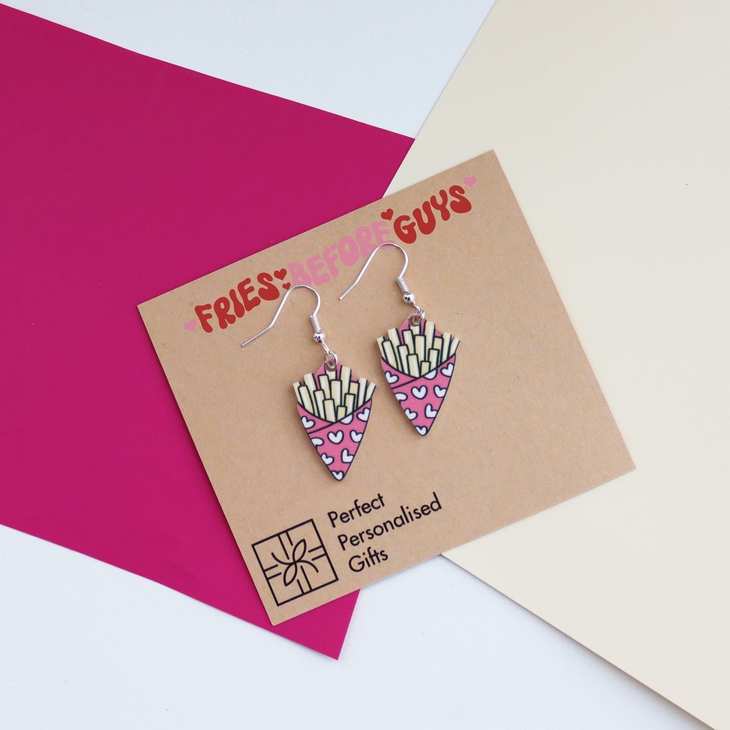 fries before guys earrings cut from acrylic and printed with a bag of chips design the bag of chips is pink and has hearts printed on it shown with an arty background