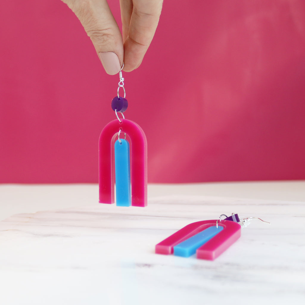 modern bright and colourful geometric arch dangle earrings cut from a purple, pink and turquoise acrylic shown hanging from a woman's fingers